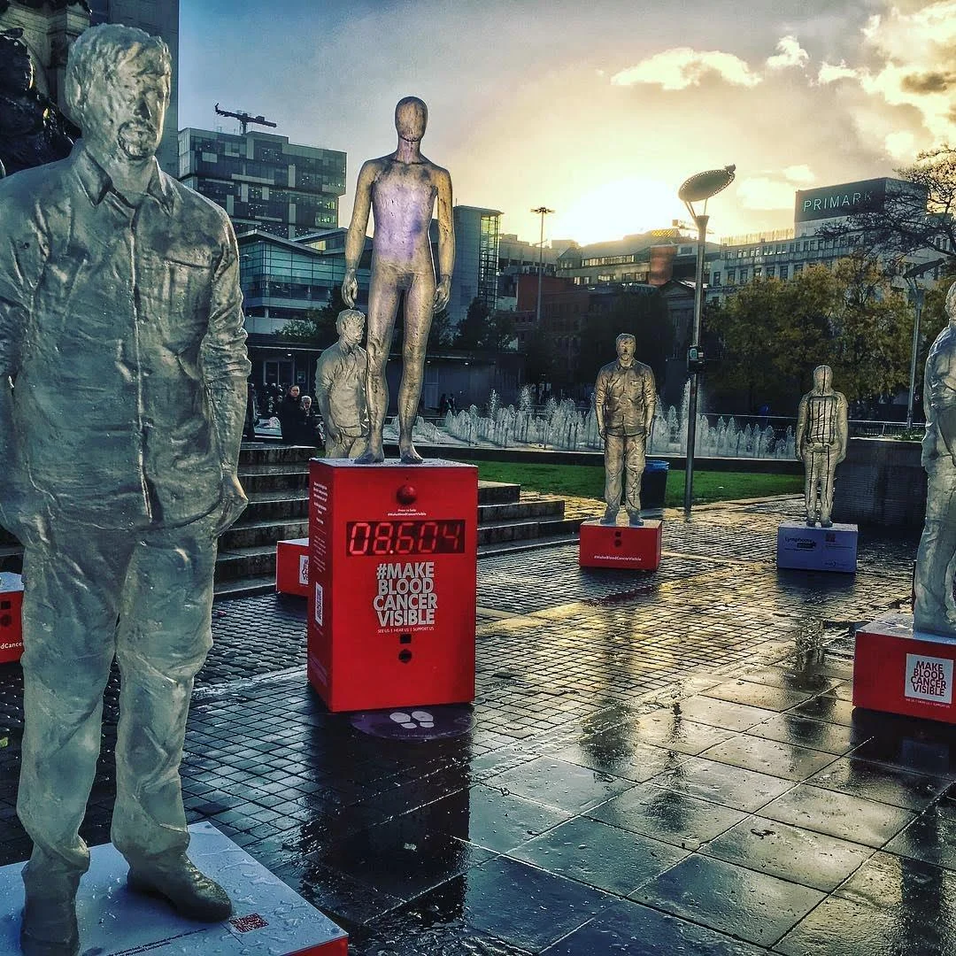 Transparent human figures on red plinths in an outdoor public square at dusk.