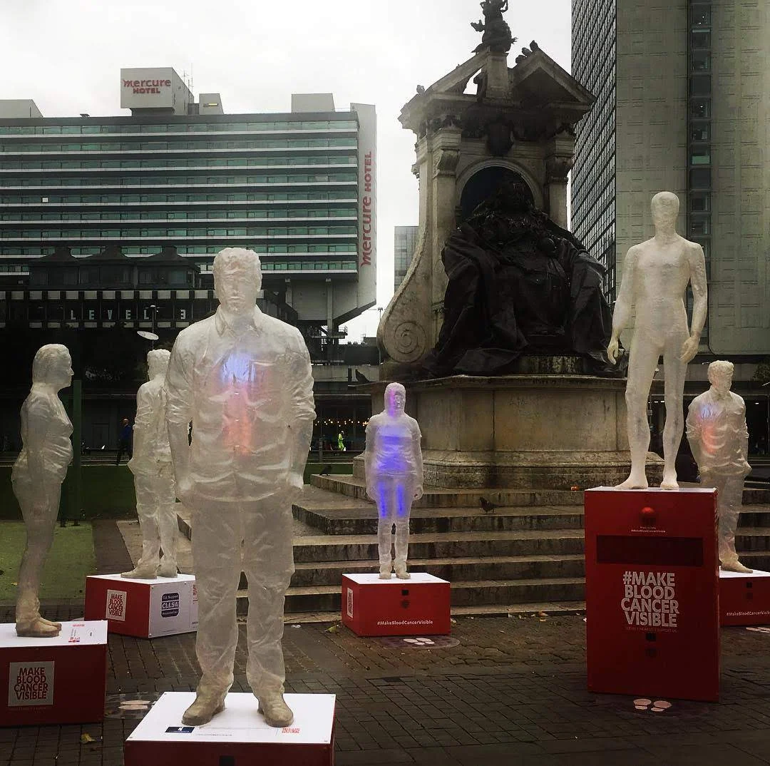Group of translucent figures on plinths beside a city monument for Make Blood Cancer Visible.