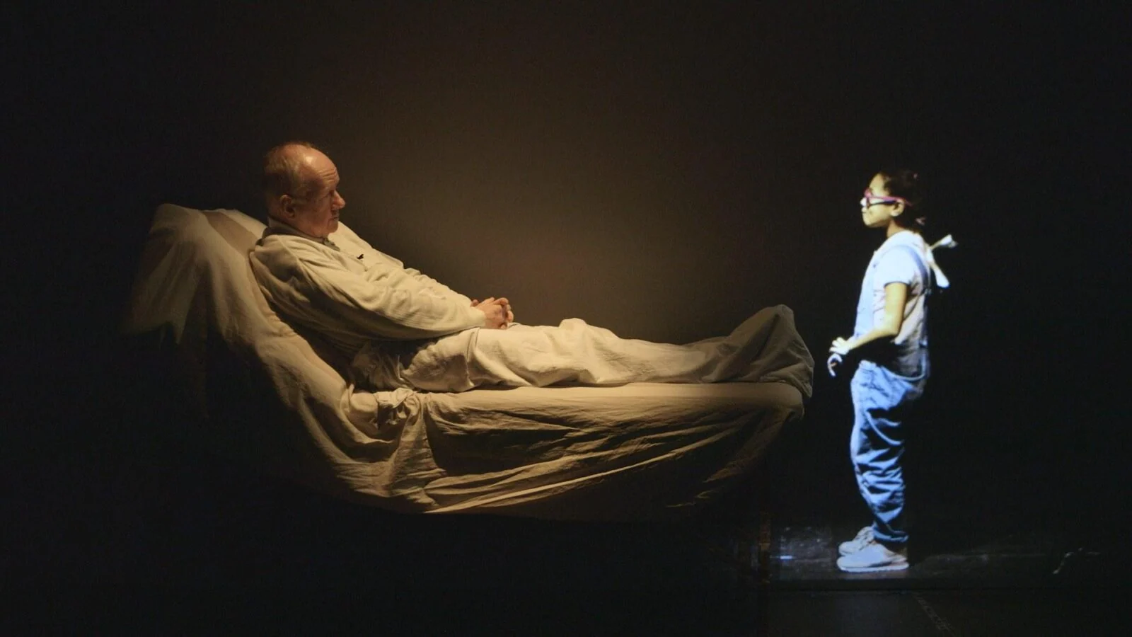 Wide view of a performer on a bed facing a projected figure in The Fall.